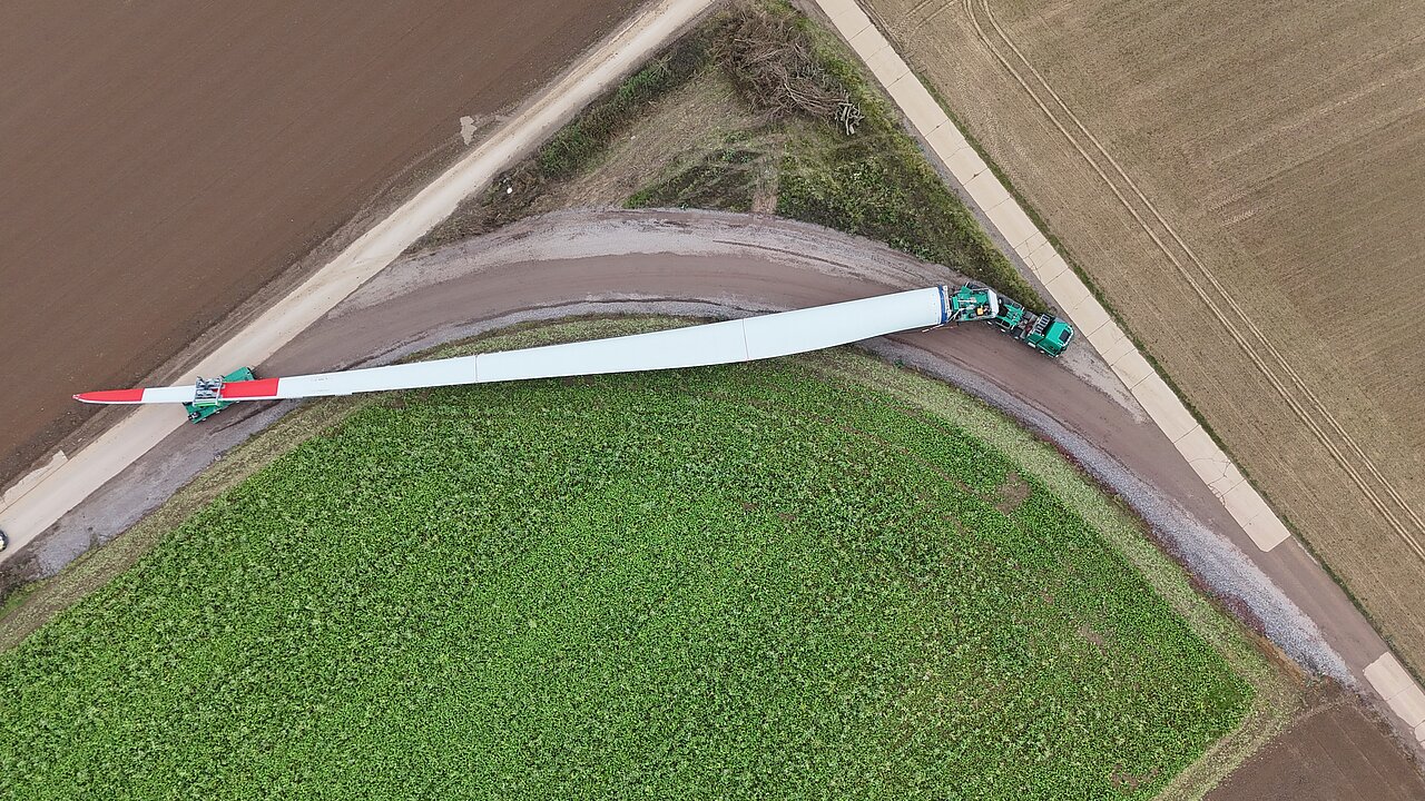 Mit Blick aus der Luft wird ein Rotorblatt mit Hilfe eines Anhängers transportiert. 