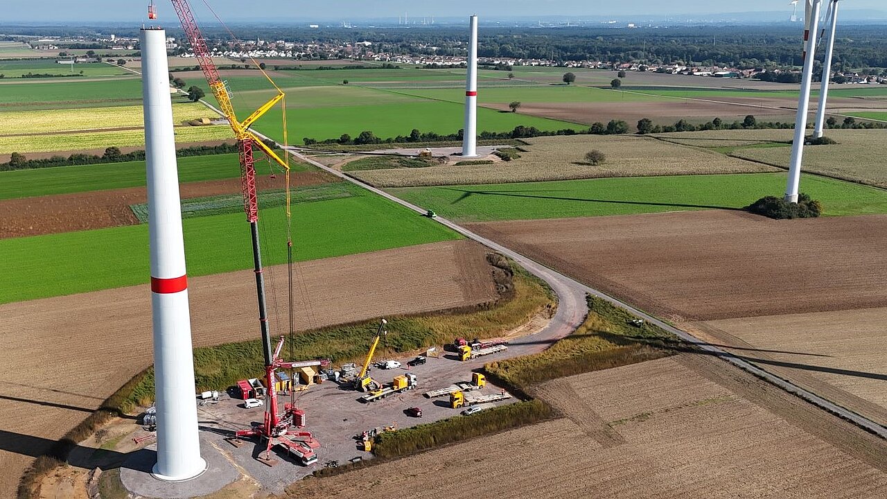 Im Vordergrund wird an einer Windenergie-Anlage gebaut. Ein Kran bringt die einzelnen Betonturmteile an.