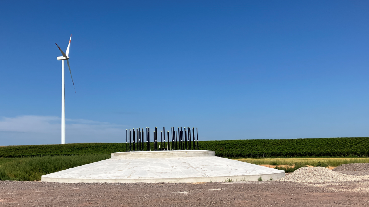 Das Fundament für eine Windenergie-Anlage steht, Im Hintergrund sind Felder und eine fertige Windenergie-Anlage.