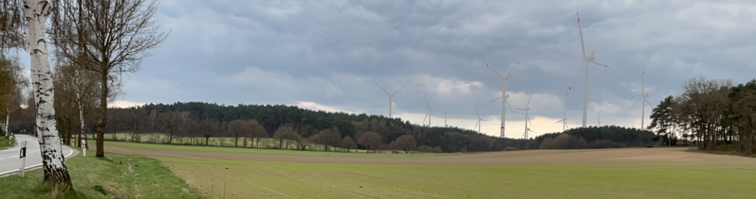 Links sieht man ein kleines Stück der K8. Rechts ein gepflügter Acker im Vordergrund. Im Hintergrund ein Nadelwald mit Windenergieanlagen. 