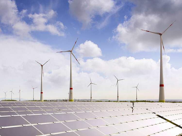 Im Vordergrrund stehen PV-Module, dahinter sind mehrere Windenergie-Anlagen zusehen. 